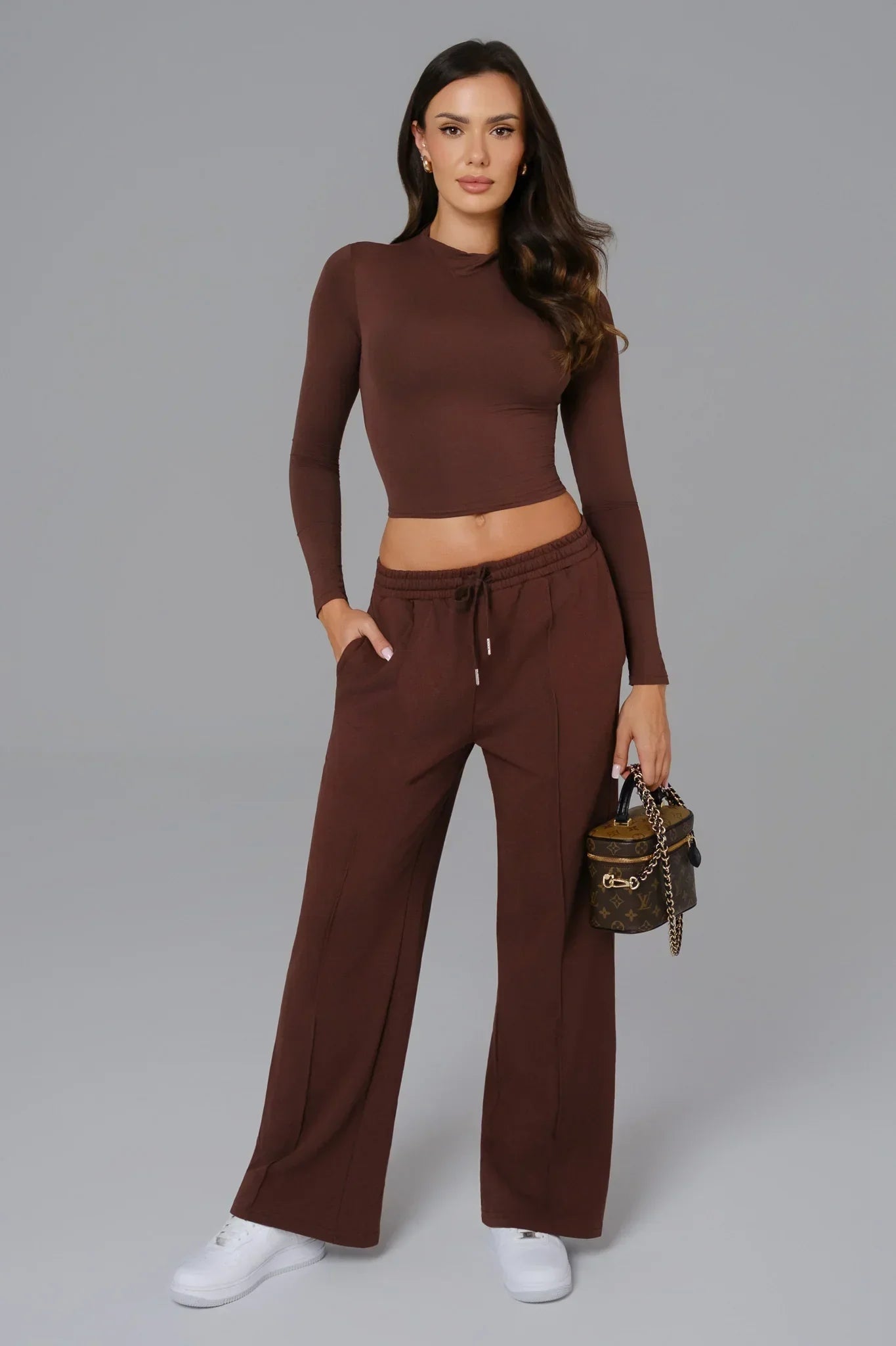 Woman wearing a brown outfit with wide-leg pants and a long-sleeve top, holding a handbag on a gray background.